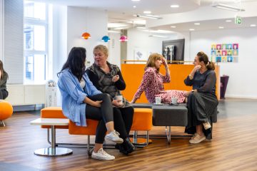 A group of adult females sat in a orange break-out area with coffee mugs