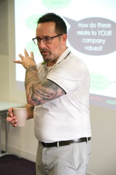 An adult male standing and holding a coffee cup while presenting at a training course event