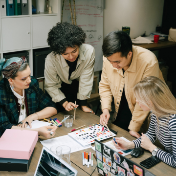 Four people in a workplace meeting and planning around a desk with a planner chart and laptop